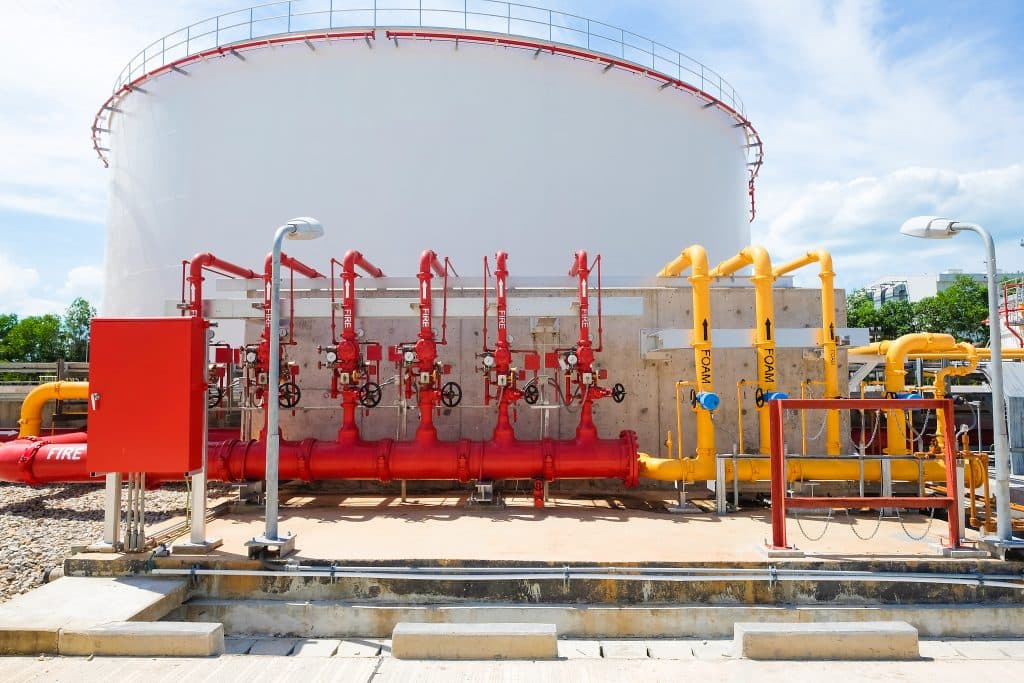Large white industrial tank with red fire suppression and yellow foam pipes in foreground.